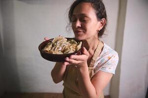 Young woman housewife in beige kitchen apron, holding a clay dish with freshly cooked homemade dumplings Varenyky with mashed potatoes and roasted onions. Traditional Ukrainian cuisine and culture photo