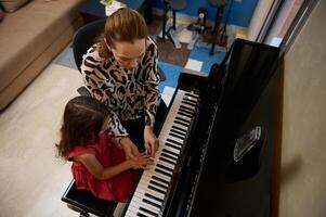 Caucasian pleasant mature female music teacher, pianist teaching piano, explaining to a little kid girl student the correct position of fingers on the piano keys. View from above photo