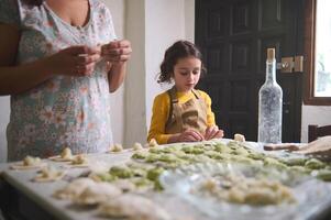 encantador niño niña en beige cocinero delantal, moldura empanadillas con machacado patata relleno, en pie a enharinado mesa cerca su madre, Ayudar su en cocina, aprendizaje Cocinando a rústico hogar cocina interior foto