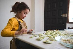 linda bebé niña en beige del chef delantal, Relleno arrollado masa rondas, haciendo empanadillas en el rural casa cocina foto