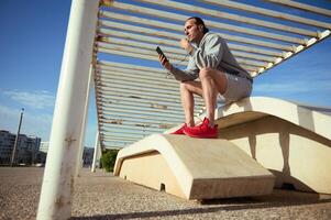 joven hermoso blanco hombre utilizando inteligente móvil teléfono mientras teniendo ejercicio descanso en el al aire libre urbano Deportes suelo foto