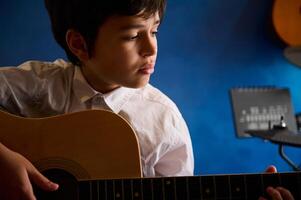 Hispano Adolescente chico jugando acústico guitarra a hogar, sentado en contra azul pared con musical instrumentos foto