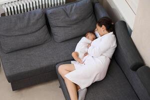 Top view young woman in white waffle bathrobe, relaxing on the couch, near her newborn baby suckling the mother's milk photo