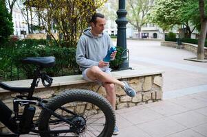 joven hombre ciclista Bebiendo agua desde Deportes botella en el ciudad, relajante después montando un Respetuoso del medio ambiente eléctrico bicicleta foto