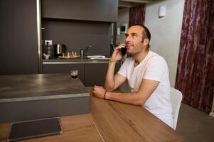 Smiling man talking on mobile phone, sitting at table at home kitchen photo