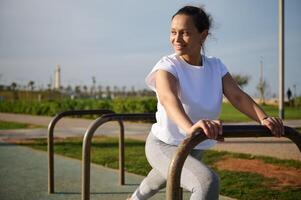 determinado Deportes mujer en blanco camiseta y gris polainas, calentamiento arriba, haciendo estocadas para extensión pierna músculos. foto