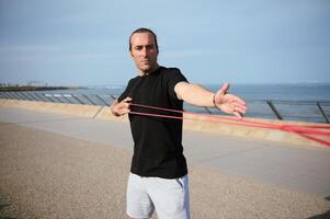 Young athlete doing arms, biceps and shoulder exercises with resistance fitness rubber bands on the beach outdoors photo