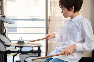 Authentic portrait of Hispanic teenager boy using drumsticks, beating on the drum while performing sounds, feeling the rhythm of music. Drum set - percussion musical instrument. photo