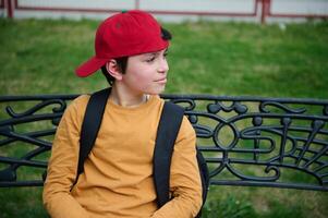 retrato de un contento Adolescente colegial en un rojo gorra, sentado en un parque banco, sonriente y mirando soñando a el lado foto