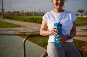 Cropped view of smiling sportswoman holding a bottle of water, relaxing after sports training on the urban sportsground photo