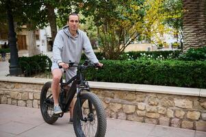 lleno longitud retrato de un hermoso caucásico hombre alquiler eléctrico bicicleta, montando en el ciudad. bicicleta compartiendo ciudad Servicio foto