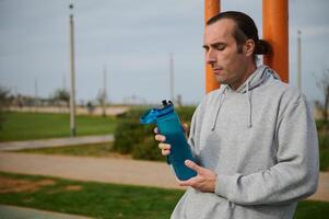 atlético hombre participación un botella de isotónico agua, descansando después pesado rutina de ejercicio en el urbano campo deportivo. personas y deporte foto