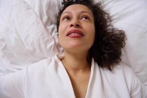 View from above smiling young adult woman waking up in the morning , lying on the bed at home bedchamber. photo