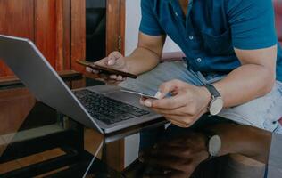 Close-up of man's hand holding cellphone and credit card. Man working using laptop at desk in home office. photo