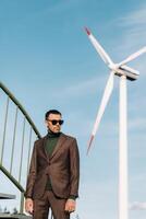 A man in a business suit with a green Golf shirt stands next to a windmill against the background of the field and the blue sky.Businessman near the windmills.Modern concept of the future. photo