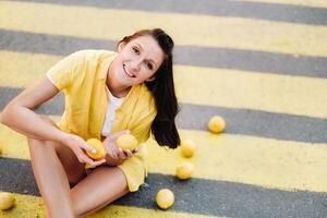 a girl holding lemons in her hands dressed in a yellow shirt, shorts and sitting on a yellow pedestrian crossing in the city and smiling. The lemon mood photo