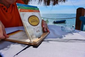 Durres, Albania - September 3rd 2022. Restaurant 2 Kitarrat Arome Deti. Man holding and reading the menu card, choosing the meal. Beautiful restaurant on Adriatic sea beach on a sunny day. photo