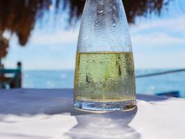 Fresh fruit water in a glass pitcher on the table in the restaurant at the sea shore. Ice cold lemonade at the beach. Detail of the bottle. Drops of water. Blue sky and sea on the background. Close-up photo