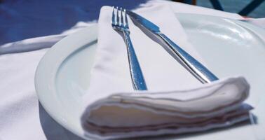 Table setting with white ceramic plate and white table cloth. Dining out in the fancy restaurant. Beautiful hot summer day. Closeup. Side view. Selective focus photo