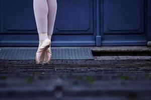 de cerca de un bailarín piernas equilibrio en el pointe zapatos. bailarina vistiendo ballet zapatillas mientras en pie en su consejos en el guijarro la carretera. gris retro puertas en el antecedentes. selectivo atención foto