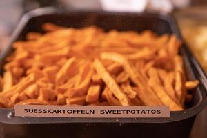 Yummy French fries in the metal tray with the tag Suesskartoffeln Sweetpotatos. Potato chips on the counter at the street market. Product Sign at market. Selective focus. Side view. Bokeh. photo