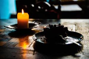 Burning wax candle and plate with a piece of berry chocolate cake and a fork  on the wooden chessboard table. Cozy evening atmosphere in cafe, coffee shop. Dark blurred background. Selective focus. photo