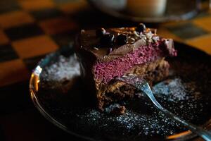 Piece of delicious homemade chocolate cake with blackberries and raspberries dusted with sugar powder served on the dark plate with fork on the wooden chess board table. Tasty Pastry ready for eating. photo