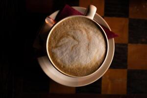 de cerca de el taza de capuchino en el blanco plato en el oscuro de madera mesa con ajedrez tablero modelo. blanco taza de caliente café latté en el tablero de ajedrez. parte superior vista. Copiar espacio. foto