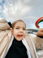 pequeño niña con su lengua colgando fuera y un caramelo en su mano en contra el fondo de un arco iris foto