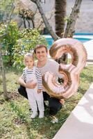Smiling dad hugging little girl squatting in garden with inflatable number 3 on string photo