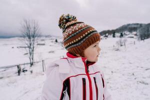 pequeño niña soportes en un cubierto de nieve colina en un montaña Valle y mira lejos foto