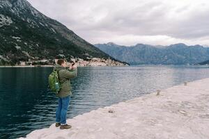 Man with a backpack stands on a pier by the sea and photographs the mountains. Back view photo