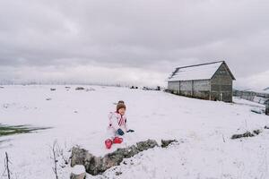 Little girl sitting on a snowy pasture near a hill farm photo