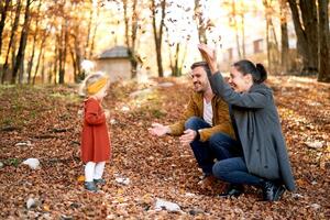 Smiling mom and dad sprinkle dry leaves on a little girl sitting on the ground in the autumn forest photo