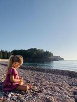 Little girl is sitting on a pebble beach by the sea and opening a small bag photo