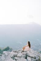 Young woman with a hat in her hand sits on a rocky cliff and looks at the mountains. Back view photo