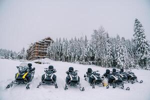 Row of snowmobiles stands on the snow at the edge of the forest on the hill of a ski resort photo