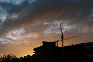siluetas de torre grúas a el construcción sitio de un de muchos pisos edificio en contra el antecedentes de un naranja puesta de sol cielo foto