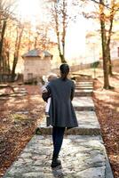 mamá con un pequeño niña en su brazos camina a lo largo un pavimentado camino en el otoño bosque. espalda ver foto