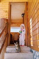 Little girl stands on the threshold of a wooden cottage and looks at a striped cat on the steps photo