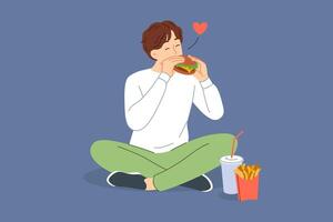 Man eats hamburger with sausage and cheese, sitting on floor and enjoying taste of fast food vector
