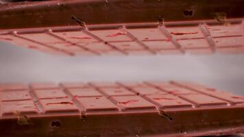 Close-up a milk ruby chocolate bar with chunks of nuts almonds and strawberry, displayed against a gray background is placed on a reflective surface, creating a mirrored image of itself video