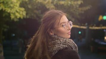 retour vue de une Jeune caucasien femme dans une manteau et écharpe tournant autour et souriant pendant une marcher dans le soir ville rue. concept de confortable et confortable l'automne Urbain la vie video