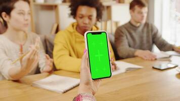 Jeune femelle employé en portant une téléphone intelligent avec une maquette vert écran proche en haut pendant une discussion de une travail projet avec multiracial entreprise équipe dans Bureau sur le commun table video