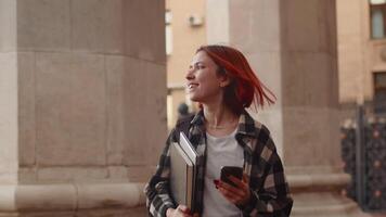 joven atractivo niña con rojo pelo vistiendo casual ropa caminando aparte el edificio con un computadora portátil, sonriente y leyendo mensajes en un teléfono inteligente video