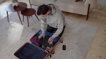 Jeune homme dans décontractée vêtements séance sur le sol à Accueil dans le confortable vivant chambre, emballage affaires dans une valise et fermeture éclair il en haut. concept de préparation à le vacances video