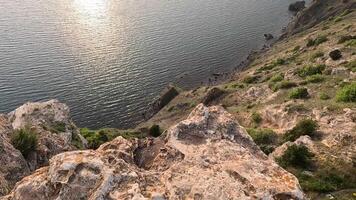 sunset over calm sea with rocky volcanic cliff. Abstract nature summer ocean sea background. Small waves on golden warm water surface with bokeh lights from sun. Slow motion video