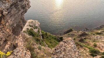 sunset over calm sea with rocky volcanic cliff. Abstract nature summer ocean sea background. Small waves on golden warm water surface with bokeh lights from sun. Slow motion video