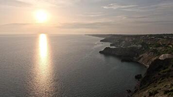 sunset over calm sea with rocky volcanic cliff. Abstract nature summer ocean sea background. Small waves on golden warm water surface with bokeh lights from sun. Slow motion video