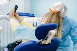 a dentist in a protective mask sits next to a patient and takes a selfie photo while working